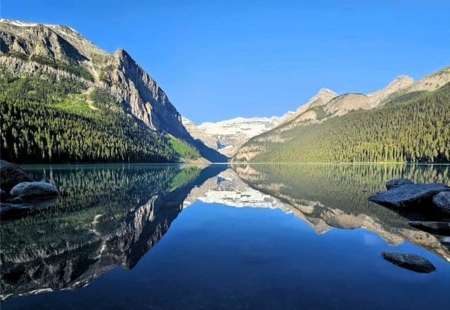Clear reflection of mountain peaks and lush forests on Lake Louise with bright blue sky and calm water.