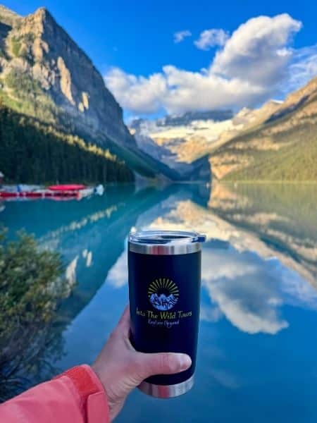 Hand holding an Into The Wild Tours mug overlooking turquoise Lake Louise with mountain and glacier reflections.