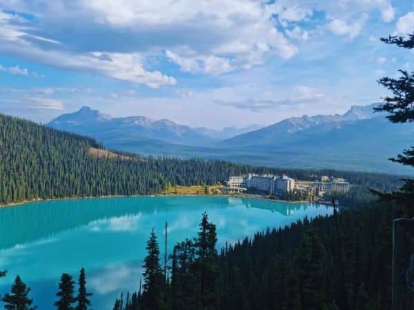 A high-angle view of turquoise Lake Louise, forested slopes, and a lakeside hotel beneath the Rocky Mountains.