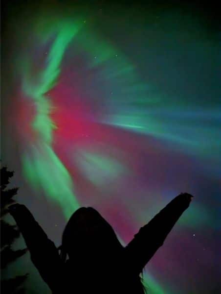 A person silhouetted against the Northern Lights over Moraine Lake, with vibrant green, pink, and purple auroras illuminating the night sky in Banff National Park.