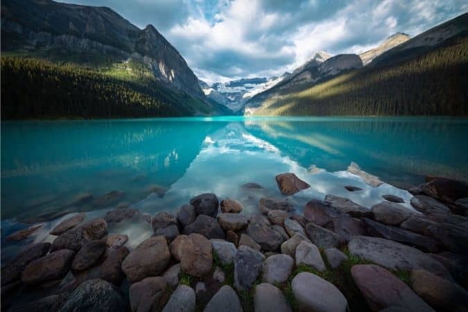 Turquoise Lake Louise with rocky shoreline, surrounded by forested mountains and glaciers under a partly cloudy sky in Banff National Park, Canada.