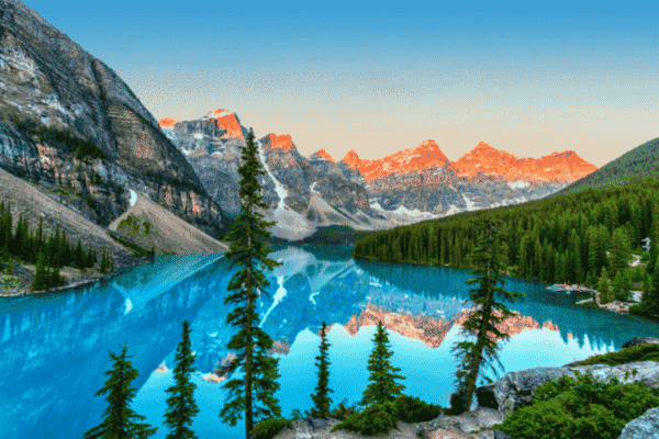 Stunning sunrise at Moraine Lake with turquoise water, pine trees, and the snow-capped Ten Peaks reflecting in the calm lake.
