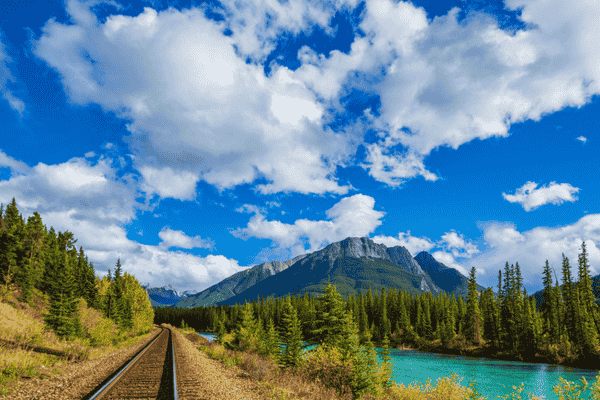 Railway tracks winding through Morant’s Curve alongside the turquoise Bow River, with pine forests and majestic Rocky Mountains under a bright blue sky.