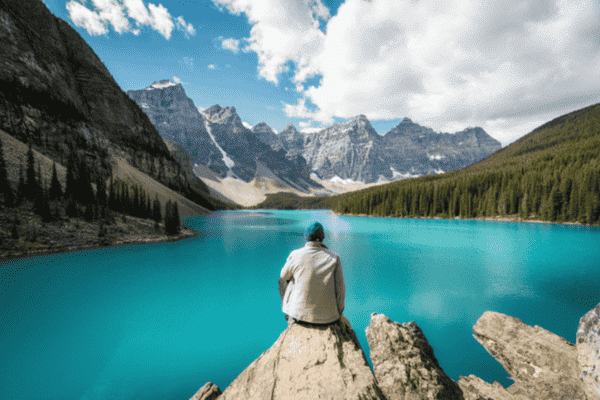 A man sitting on a rock overlooking the vivid turquoise waters of Moraine Lake, surrounded by forested valleys and towering Rocky Mountain peaks.