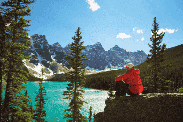 Person in a red jacket sitting on a rock, overlooking the turquoise waters of Moraine Lake surrounded by evergreen trees and the rugged Rocky Mountains.