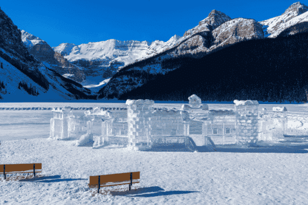 Scenic view of ice castles on frozen Lake Louise surrounded by snow-capped mountains and evergreen trees under a clear blue sky.