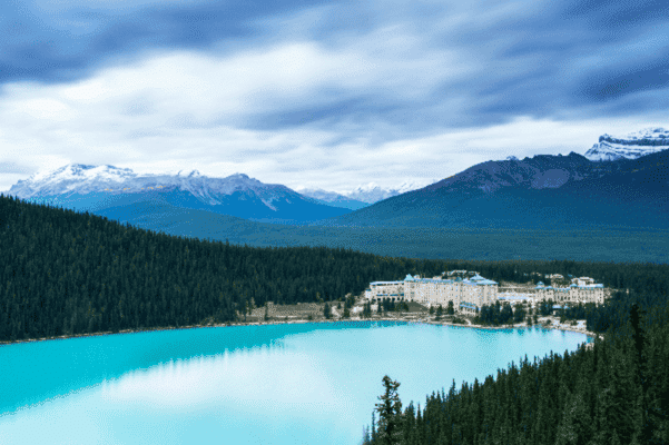 Lake Louise with turquoise water, Fairmont Chateau in the distance, and forested mountains under a cloudy sky.