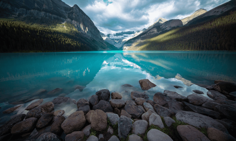 Turquoise Lake Louise with rocky shoreline, surrounded by forested mountains and glaciers under a partly cloudy sky in Banff National Park, Canada.