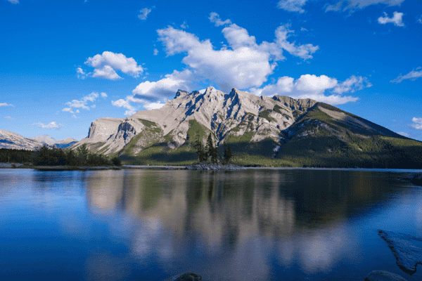 Glassy reflection of mountains at Lake Minnewanka in Banff National Park under a bright blue sky with scattered clouds.