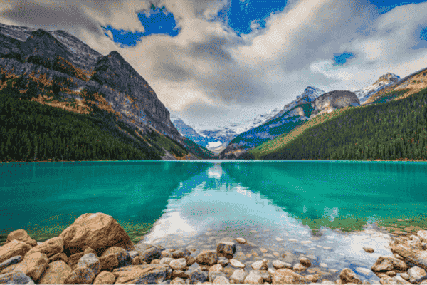 Rocky shoreline at Lake Louise with turquoise water, mountain valley, and distant glacier under morning clouds