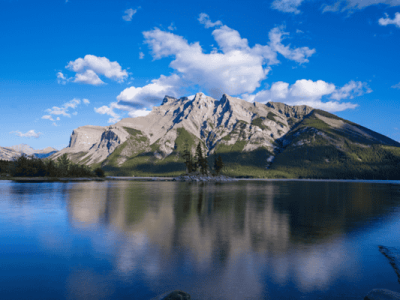Glassy reflection of mountains at Lake Minnewanka in Banff National Park under a bright blue sky with scattered clouds.