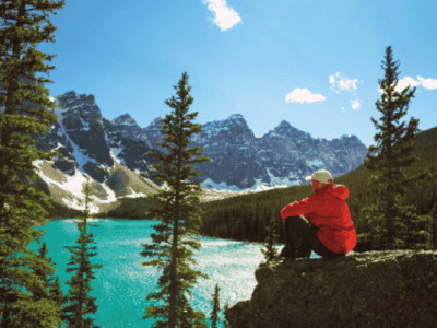 Person in a red jacket sitting on a rock, overlooking the turquoise waters of Moraine Lake surrounded by evergreen trees and the rugged Rocky Mountains.